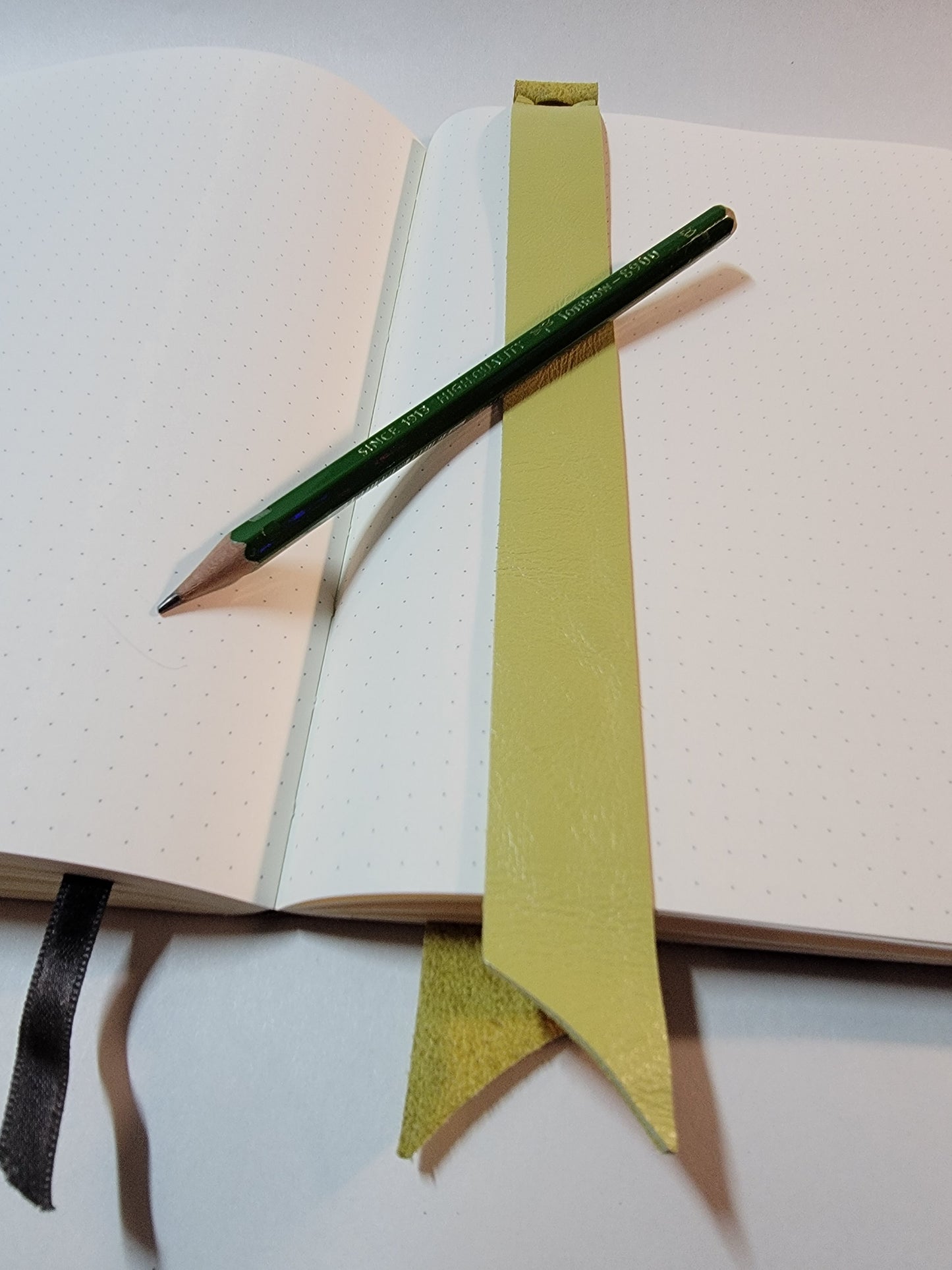 A yellow-green Sewnota Standard bookmark sitting on an open A5 dot grid notebook. A green Tombow pencil rests diagonally across the pages.