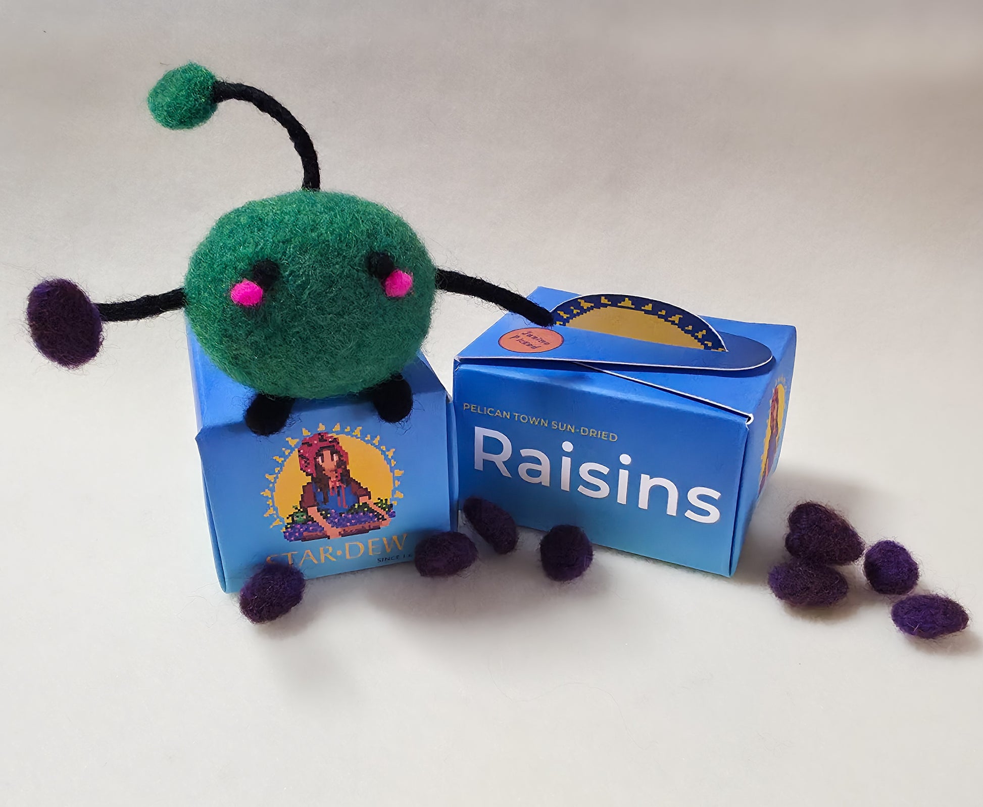 Felt toy figure of a green Junimo character  sitting on top of a blue box labelled 'Raisins'. The Junimo is holding a raisin and there are felt raisins scattered around the base of the box.