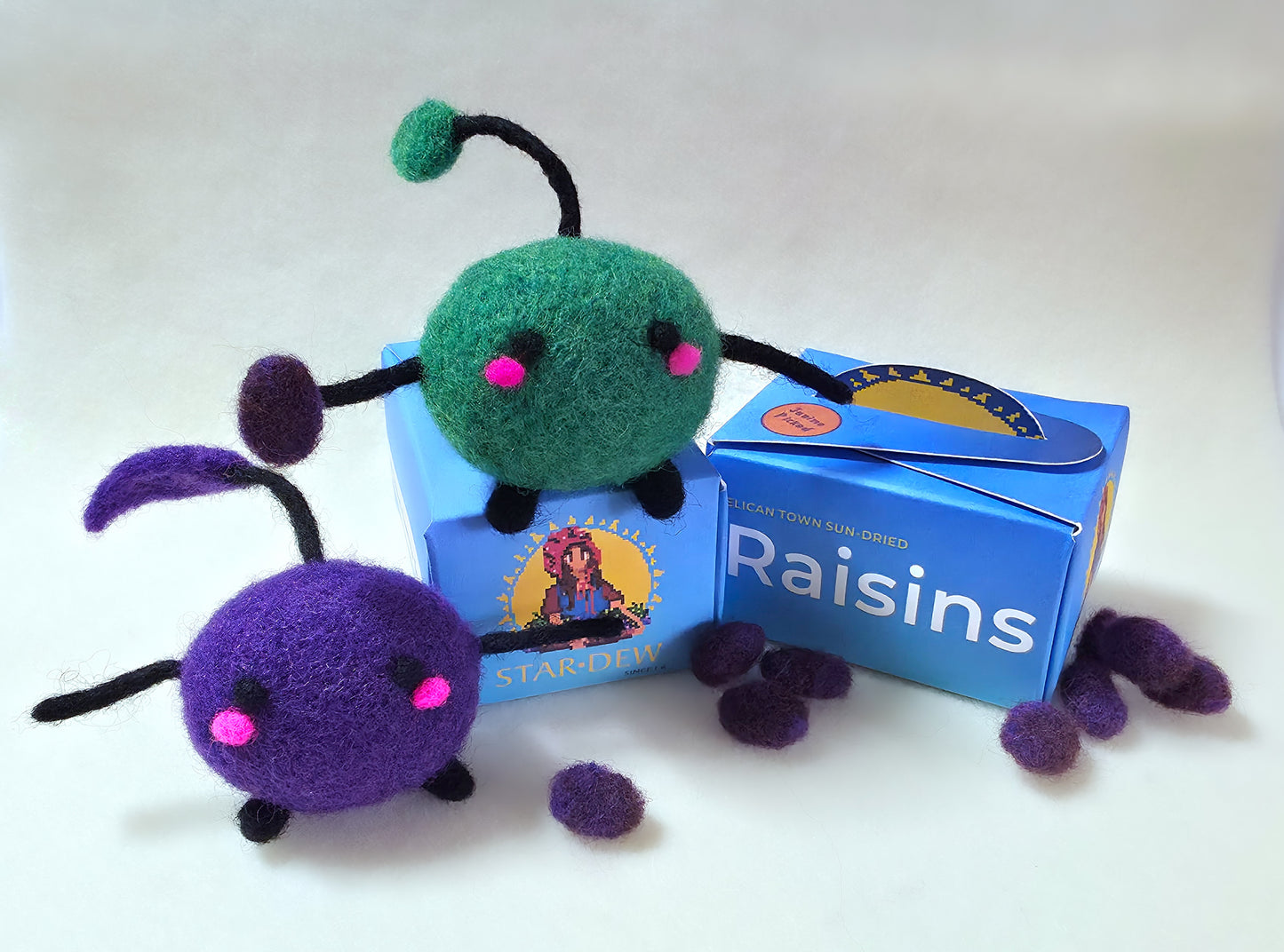 Felt toy figures of green and purple Junimo characters sitting on top of a blue box labelled 'Raisins'. The Junimo is holding a raisin and there are felt raisins scattered around the base of the box.
