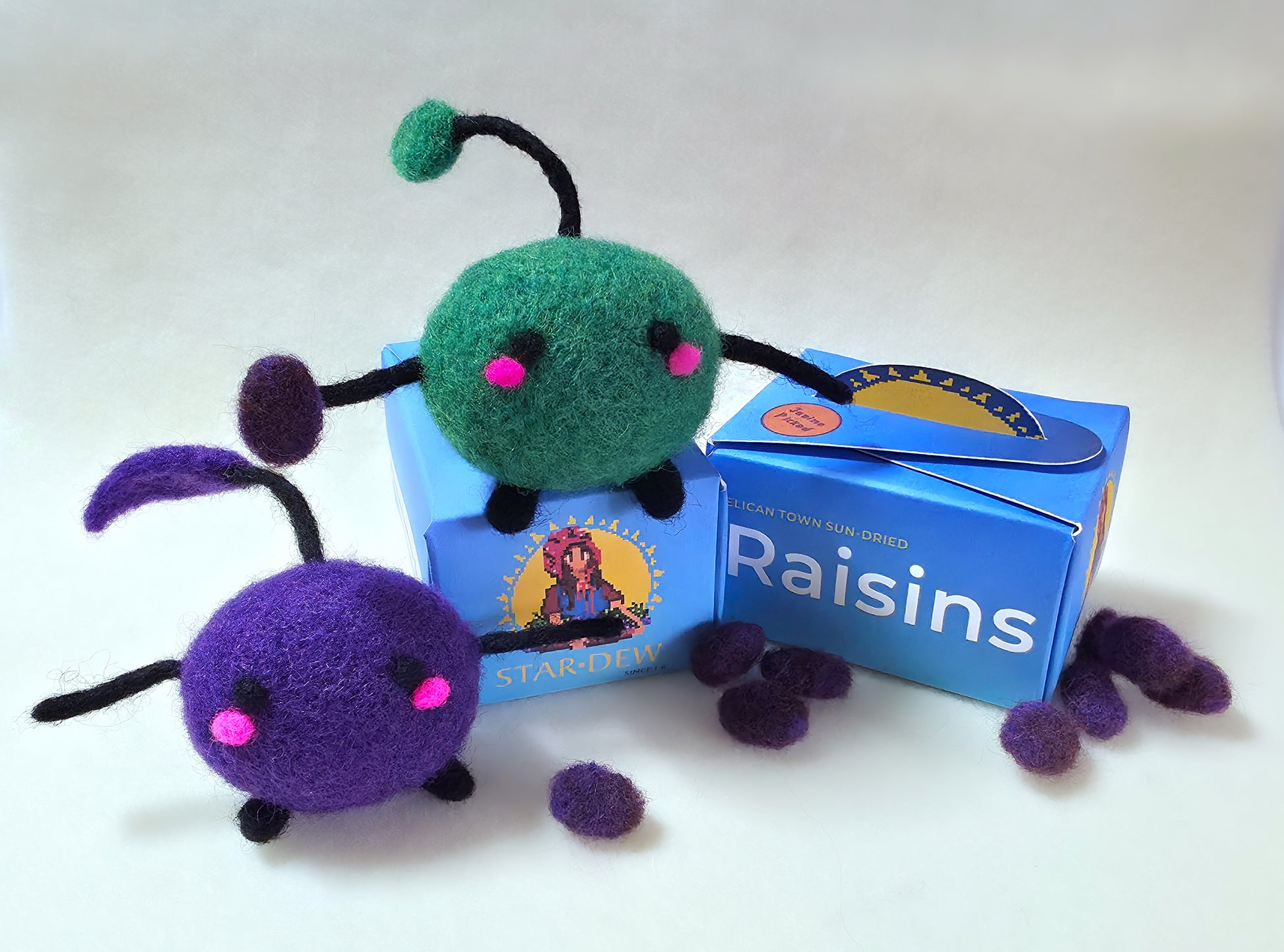 Felt toy figures of green and purple Junimo characters sitting on top of a blue box labelled 'Raisins'. The Junimo is holding a raisin and there are felt raisins scattered around the base of the box.