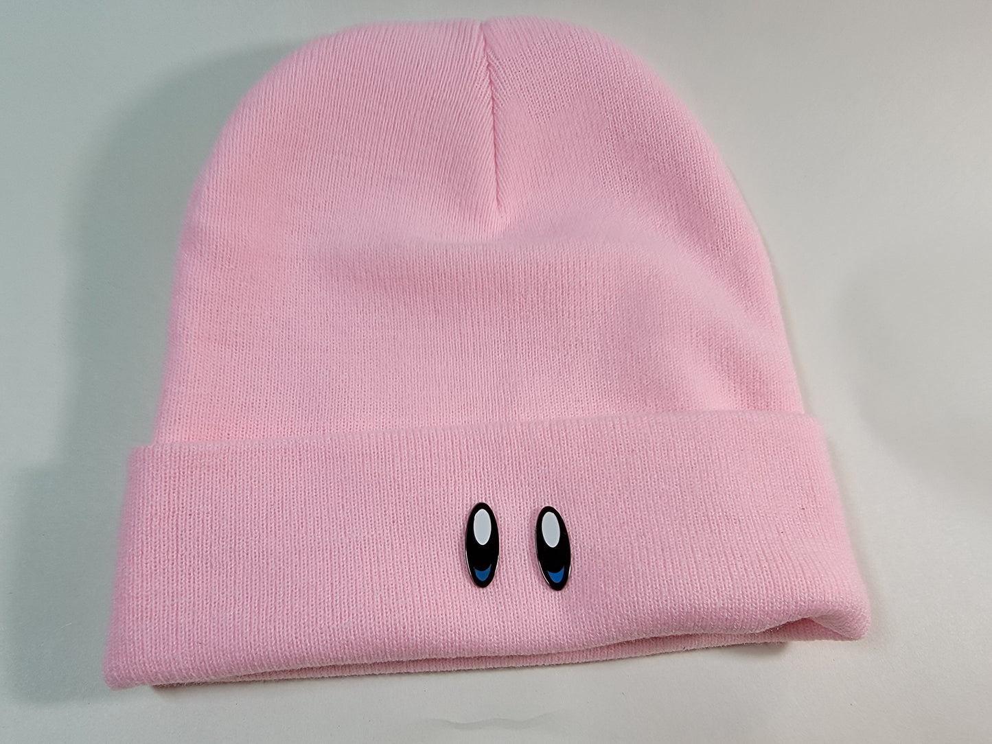 Two black metal lapel pins with white and blue enamel displayed on the bottom edge of a soft pink toque to resemble an animated characters from retro video games.