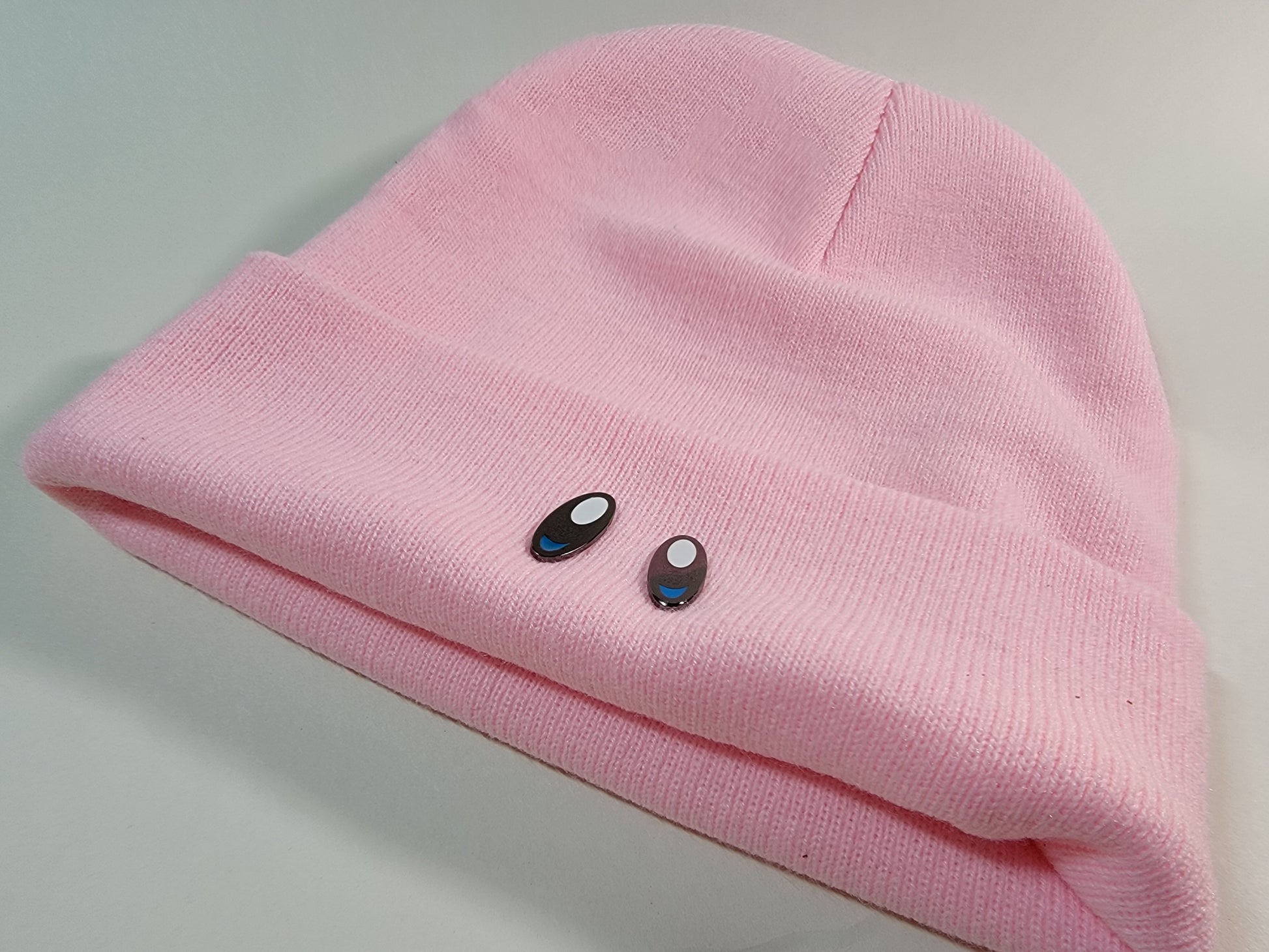 Two black metal lapel pins with white and blue enamel displayed on the bottom edge of a soft pink toque to resemble an animated characters from retro video games.