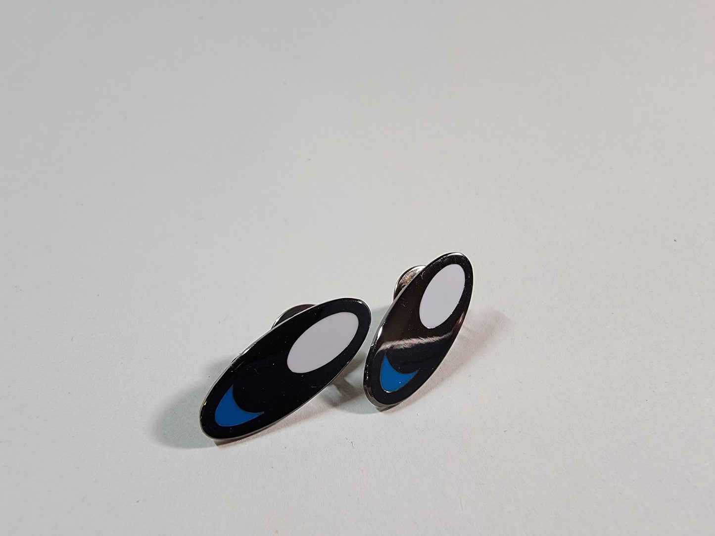 A closeup of two black metal lapel pins with white and blue enamel that resemble animated characters from retro video games.