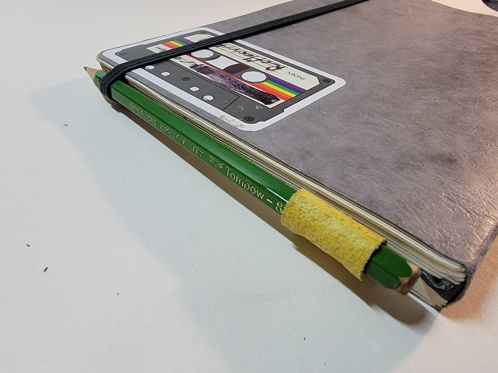 A close-up of the top of an A5 notebook. The barrel of a yellow-green Sewnota Standard bookmark is sticking out of the top of the notebook, holding a green Tombow pencil.